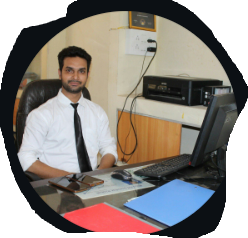 A man in a white shirt and black tie sitting at an office desk with a computer and folders, framed in a circular crop.